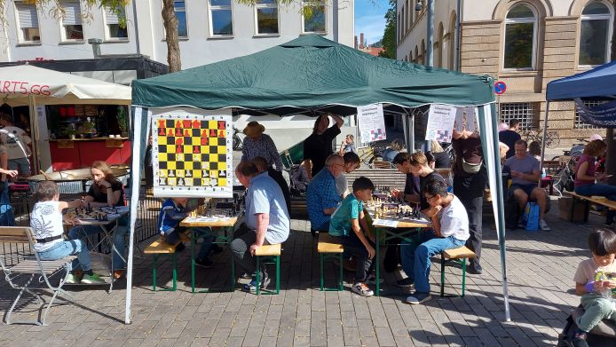 Das Bild zeigt einen grünen Pavillon, an dem Flyer und ein Demobrett hängen. An zwei Biertischen sitzen VEreinsmitglieder und Gäste beim Schach spielen. Es ist warmes, sonniges Wetter. Leute haben T-Shirts und teilweise kurze Hosen an. 