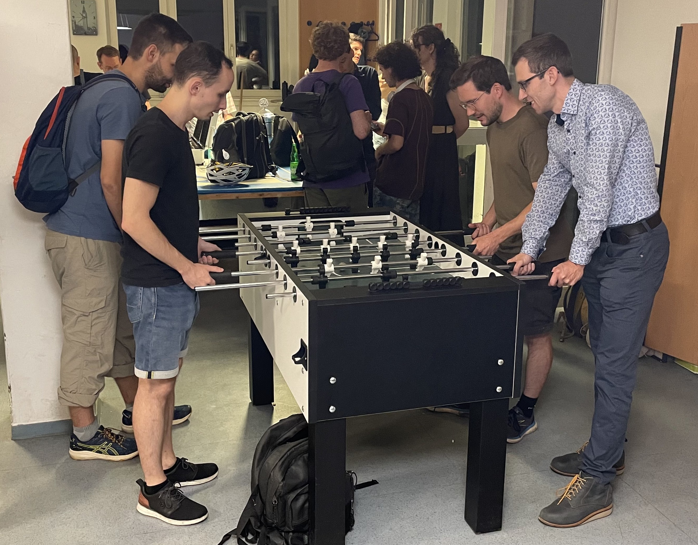 Ein Foto vom Eingangsbereich unseres Spiellokals. Im Vordergrund spielen vier Teilnehmer Fussball am Tischkicker. Im Hintergrund stehen mehrere Teilnehmer zusammen und diskutieren. Draußen ist es schon dunkel.