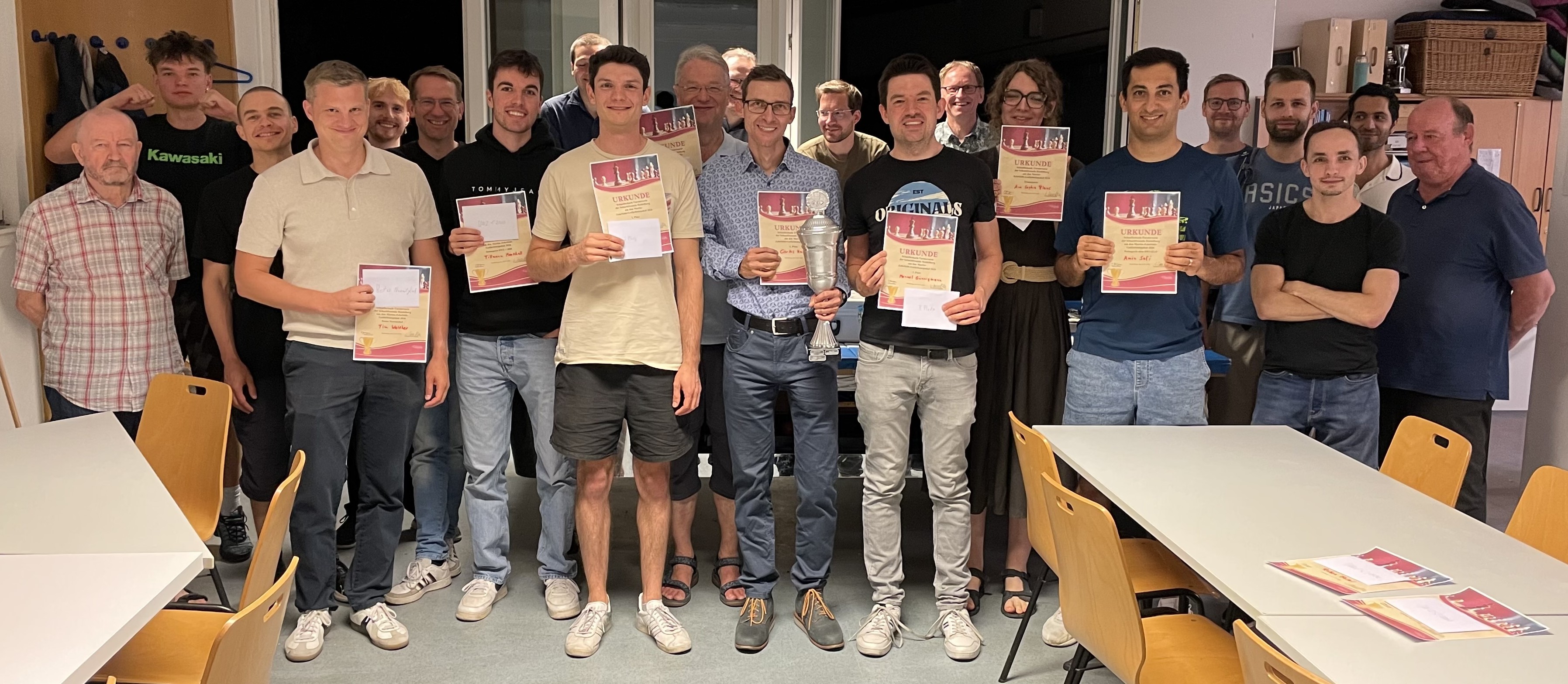 Foto im Eingangsbereich unseres Spiellokals, draußen ist es schon dunkel. Gruppenfoto mit mehr als 20 Personen. Im Zentrum stehen die Preisträger mit Urkunden und Umschlägen in der Hand. Der Sieger hält den Pokal. Drumherum stehen alle anderen Teilnehmer, die bis zum Schluss geblieben sind. Alle lächeln gut gelaunt in die Kamera.