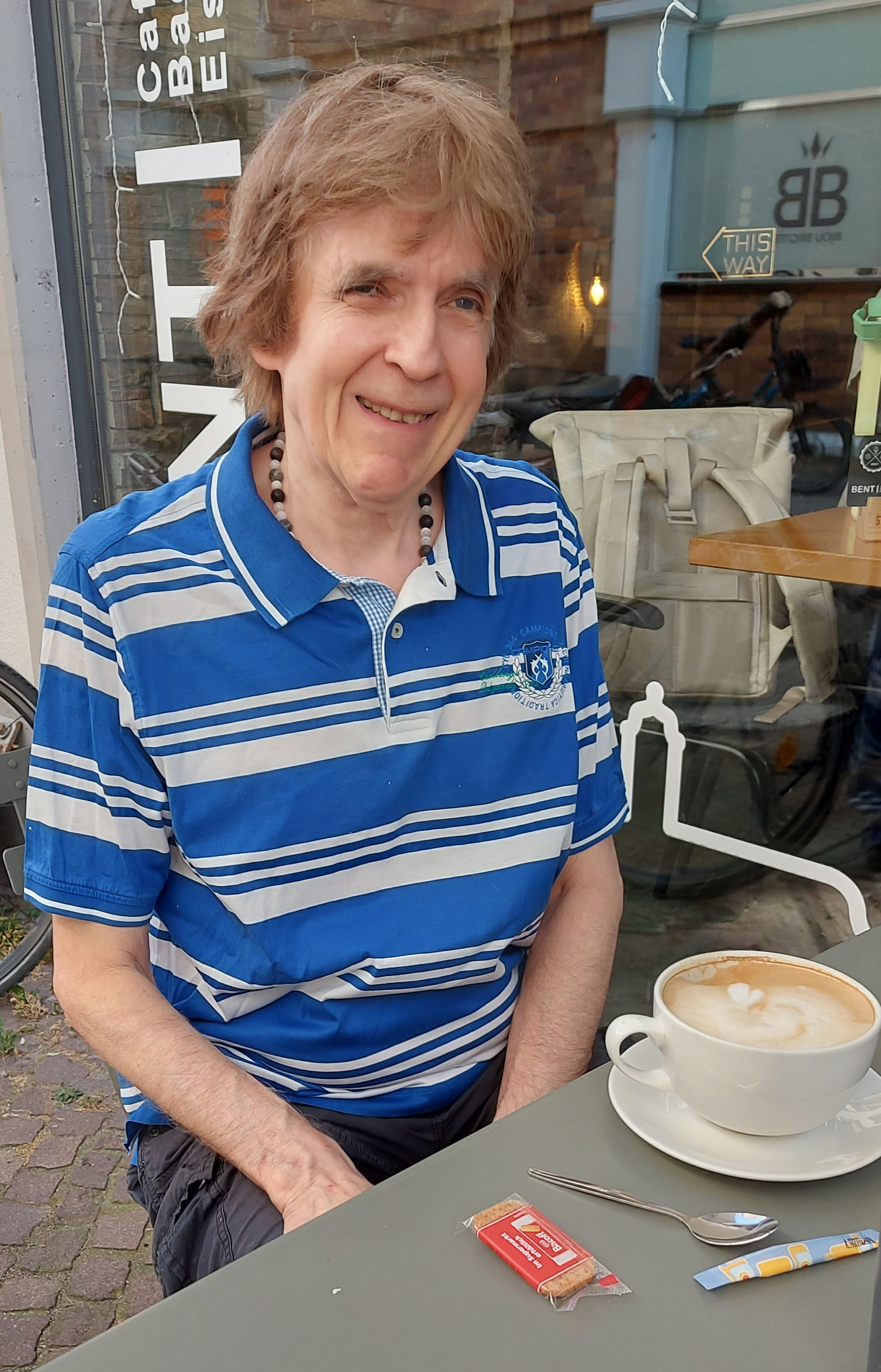 Dieter sitzt an einem grauen Blechtisch vor einem Cafe in Heidelberg. Er trägt ein blau-weiß gestreiftes Polo-Shirt und lächelt freundlich in die Kamera. Vor ihm steht eine große, volle Tasse Milchkaffee. Der Milchschaum darauf hatte bestimmt mal das Muster eines Kleeblattes, ist aber schon ziemlich zerlaufen.