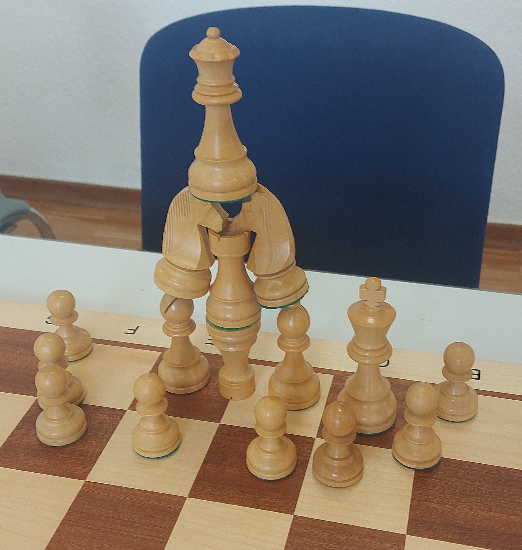 Das Foto zeigt eine Pyramide aus weißen Schachfiguren, aufgebaut auf einem Holzschachbrett. Zwei Türme stehen übereinander, links und rechts daneben steht ein Läufer. Auf den Läufern stehen Springer, die mit der Nase auf dem Turm liegen. Ganz oben auf beiden Springern steht die Dame. König und Bauern stehen neben der Pyramide. 